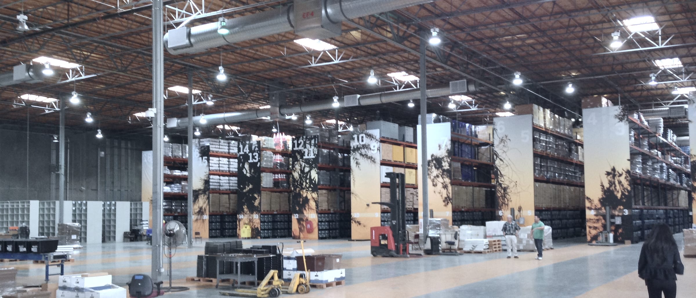 An aisle between two long rows of stored lever voting machines in a board of elections warehouse, under bare overhead lamps.