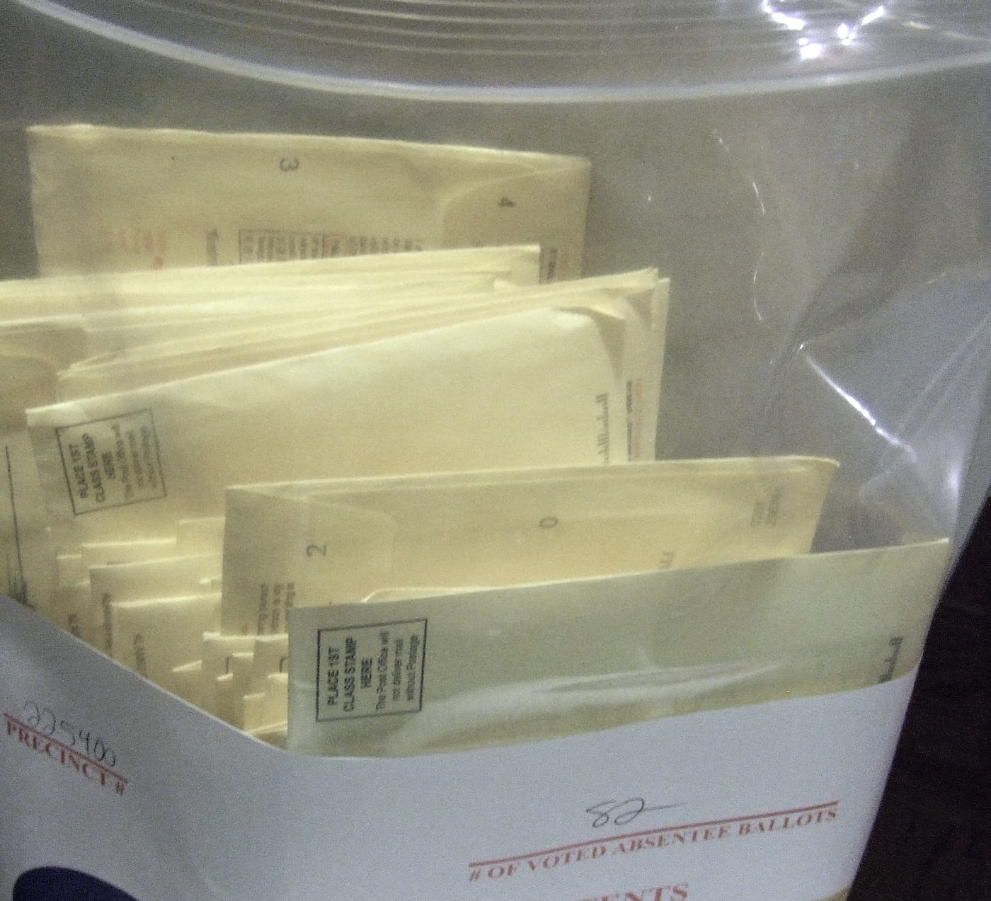 An election worker bent over a clear plastic ballot bag under a single hard light at night, one hand on the bag's label, with more filled bags stacked beside him in the dark.
