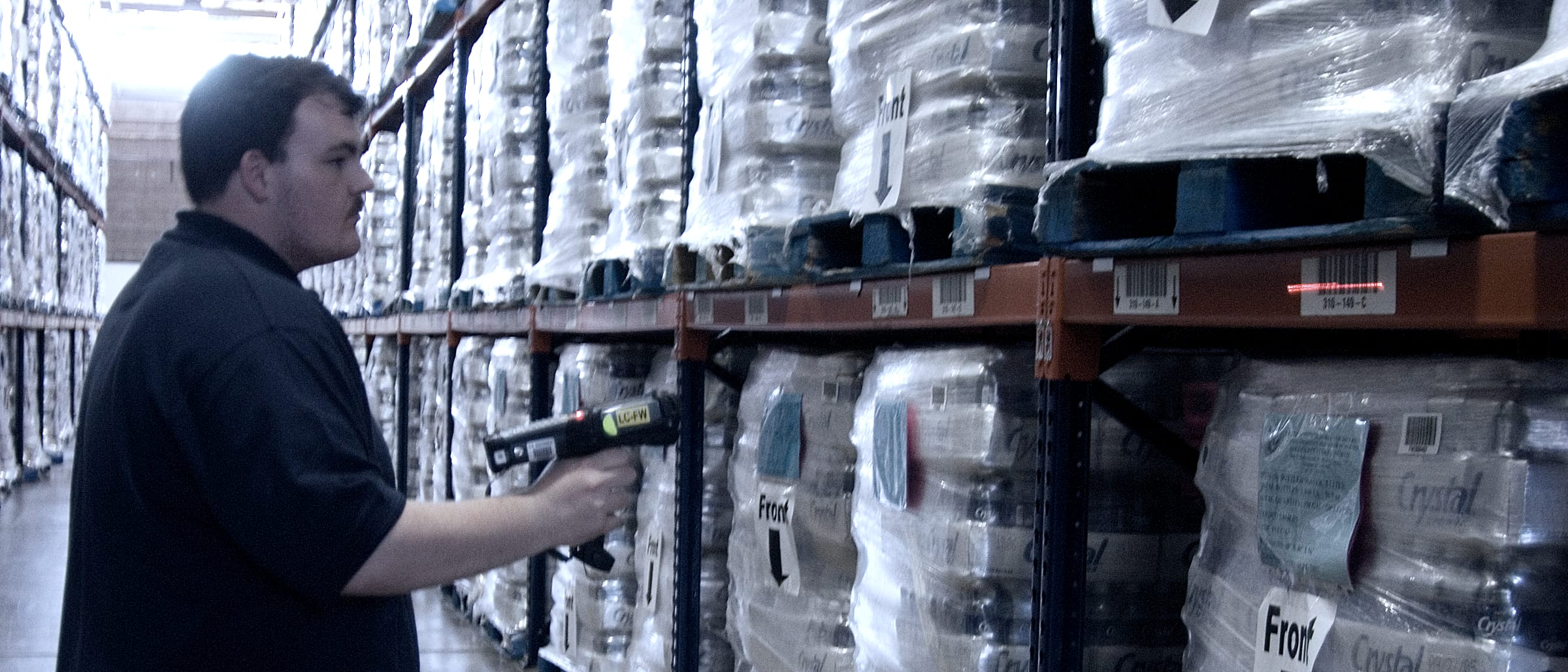 A warehouse aisle between racks of shrink-wrapped pallets. A worker aims a pistol-grip scanner at a location label on a rack beam; the red laser line falls across the label.